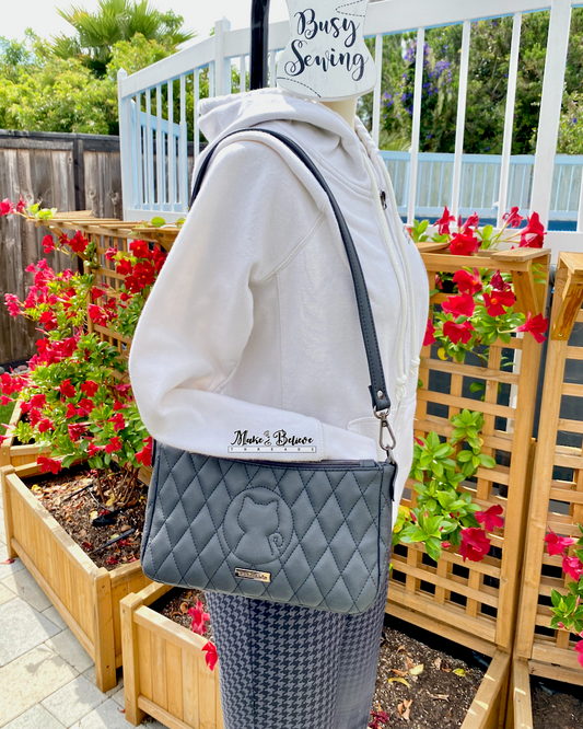Mannequin wearing a white hoodie, holding a quilted charcoal black shoulder bag with a quilted cat silhouette, standing in front of a garden with flowers.