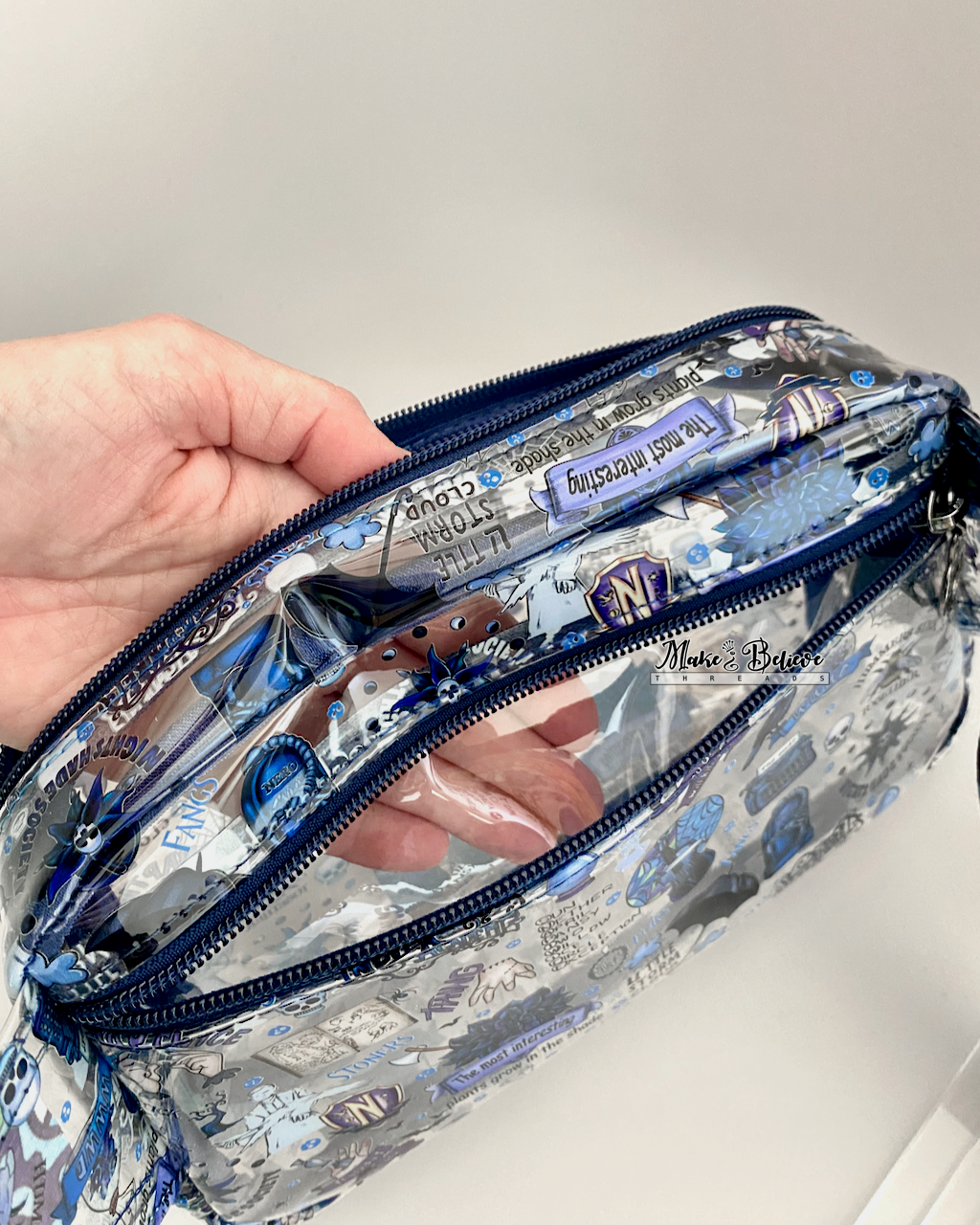 Hand inside the main zipper compartment of a clear pouch. The back zipper compartment is also open and the hand is visible through the layer that separates the two compartments. Photographed on a white background.