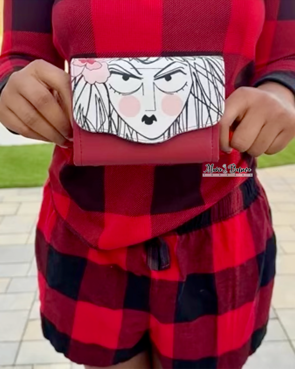 Person wearing a red and black checkered outfit holding a mini Necessary Clutch Wallet with a drawn face of a woman, print from Alexander Henry's Ghastlies collection
