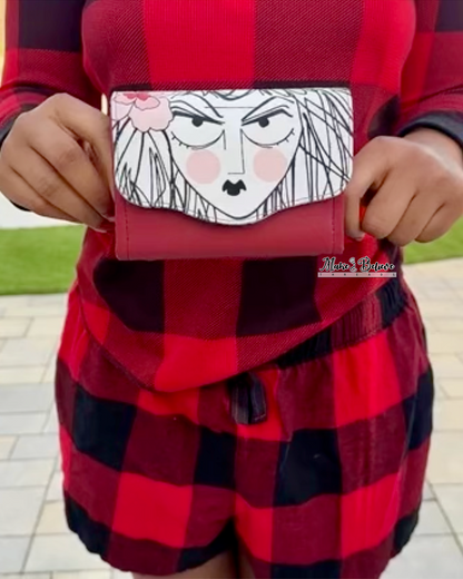Person wearing a red and black checkered outfit holding a mini Necessary Clutch Wallet with a drawn face of a woman, print from Alexander Henry's Ghastlies collection