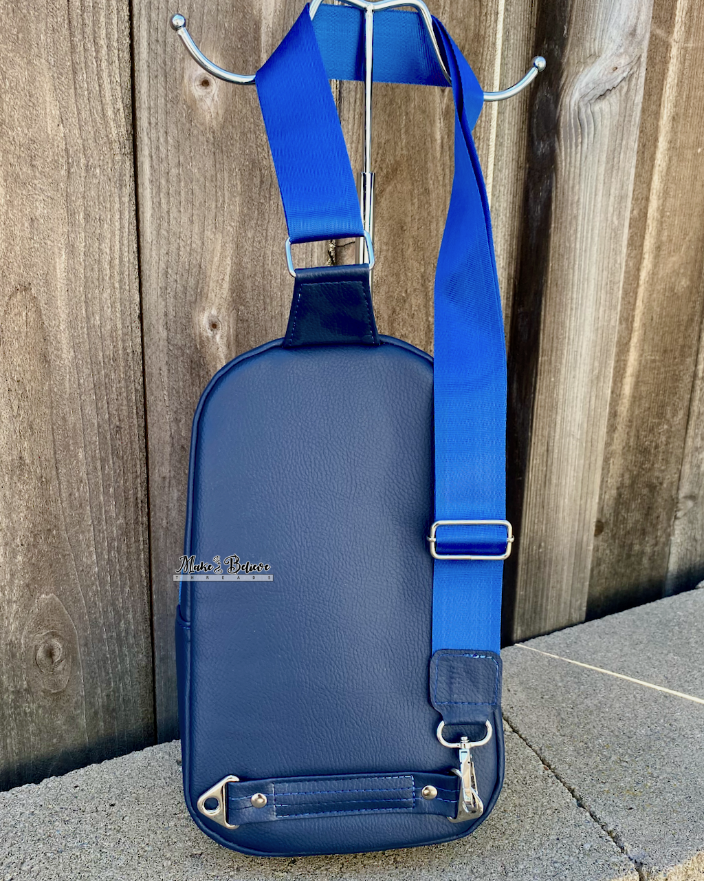 Back view of a navy sling bag with a blue strap and nickel hardware on a stone wall with wooden background