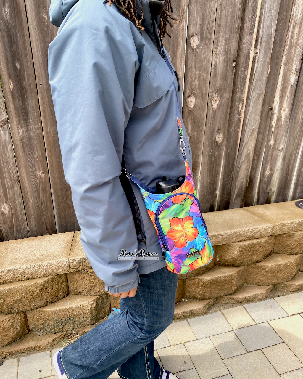 Person wearing a colorful floral water bottle carrier crossbody walking in front of a wooden fence