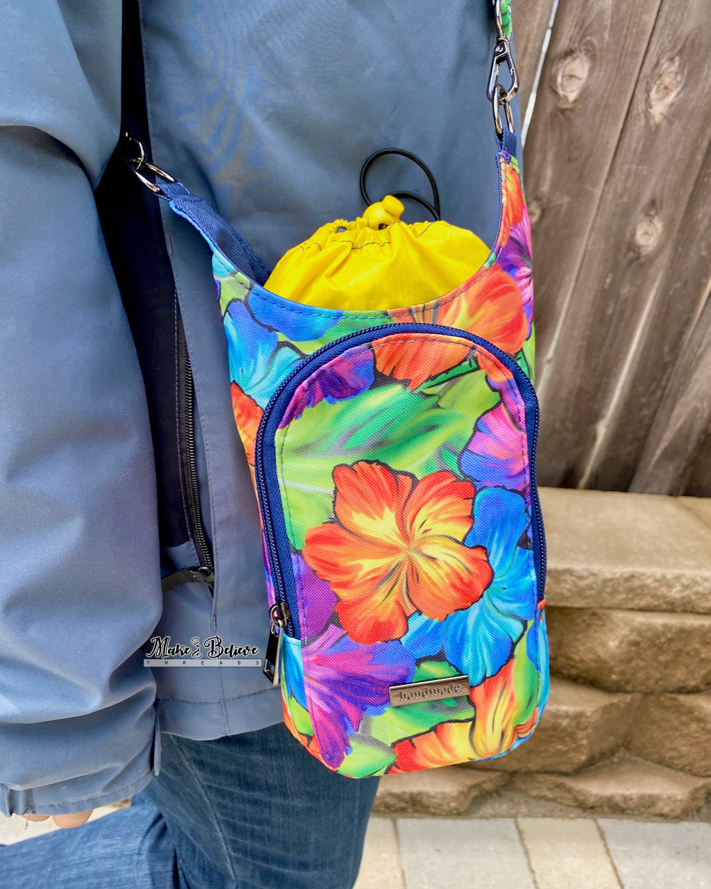 Colorful floral water bottle carrier with  a yellow drawstring lining cinched over a water bottle, photographed hanging from a person's shoulder against a wooden background.