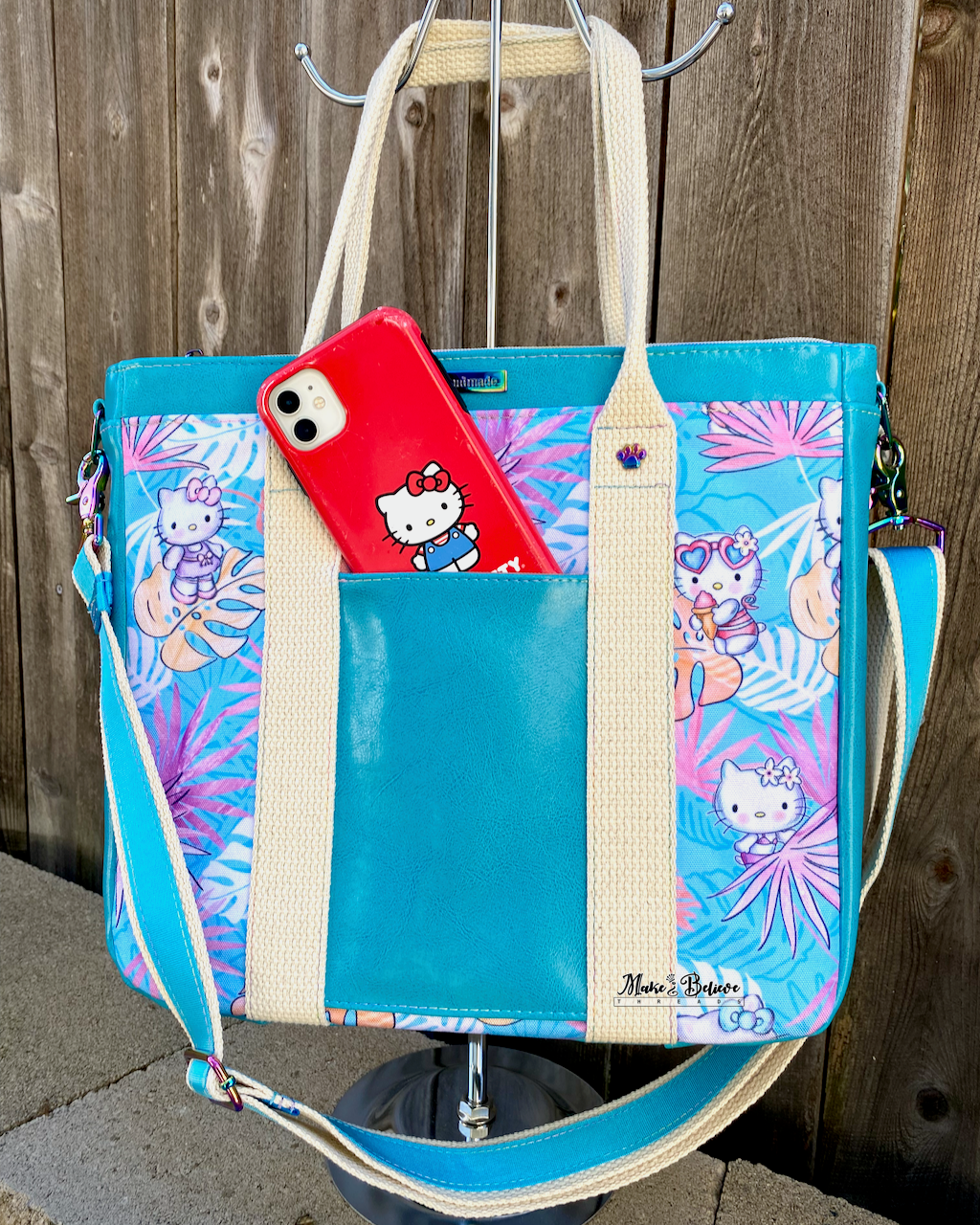 Aqua blue tote bag with pink and blue tropical print featuring "Bow Kitty" in front of a wooden fence. There is a red "Bow Kitty" phone peeking out from the from slip pocket.