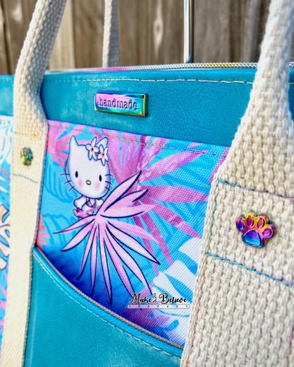 Close-up of an aqua blue tote bag with pink and blue tropical print featuring "Bow Kitty" and rainbow paw print chicago screws and handmade label, photographed in front of a wooden fence.