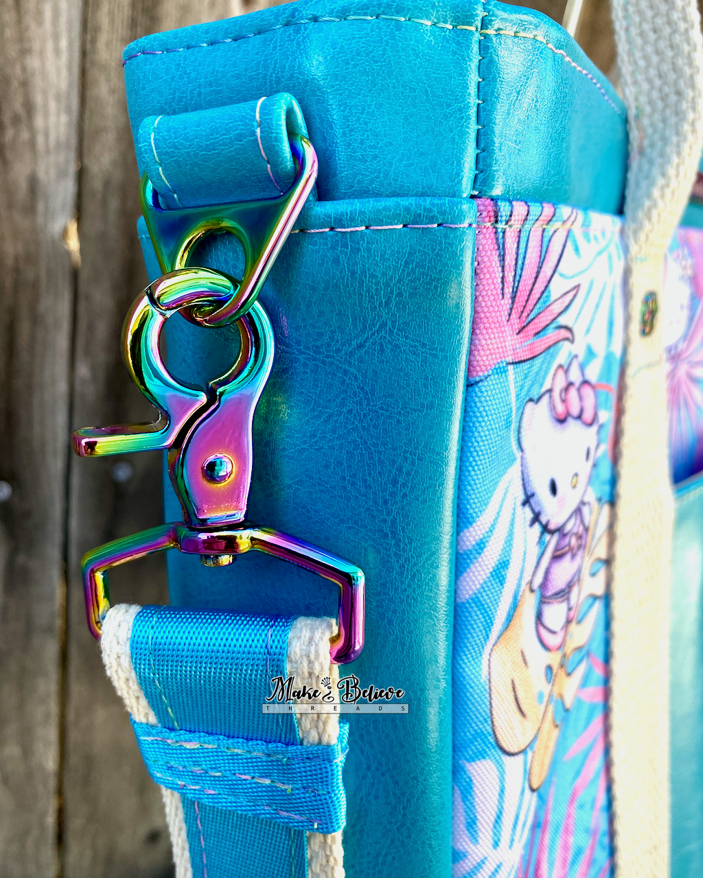 Close-up of a rainbow side connector and triangle ring on a aqua blue tote featuring "Bow Kitty," photographed in front of a wooden fence.