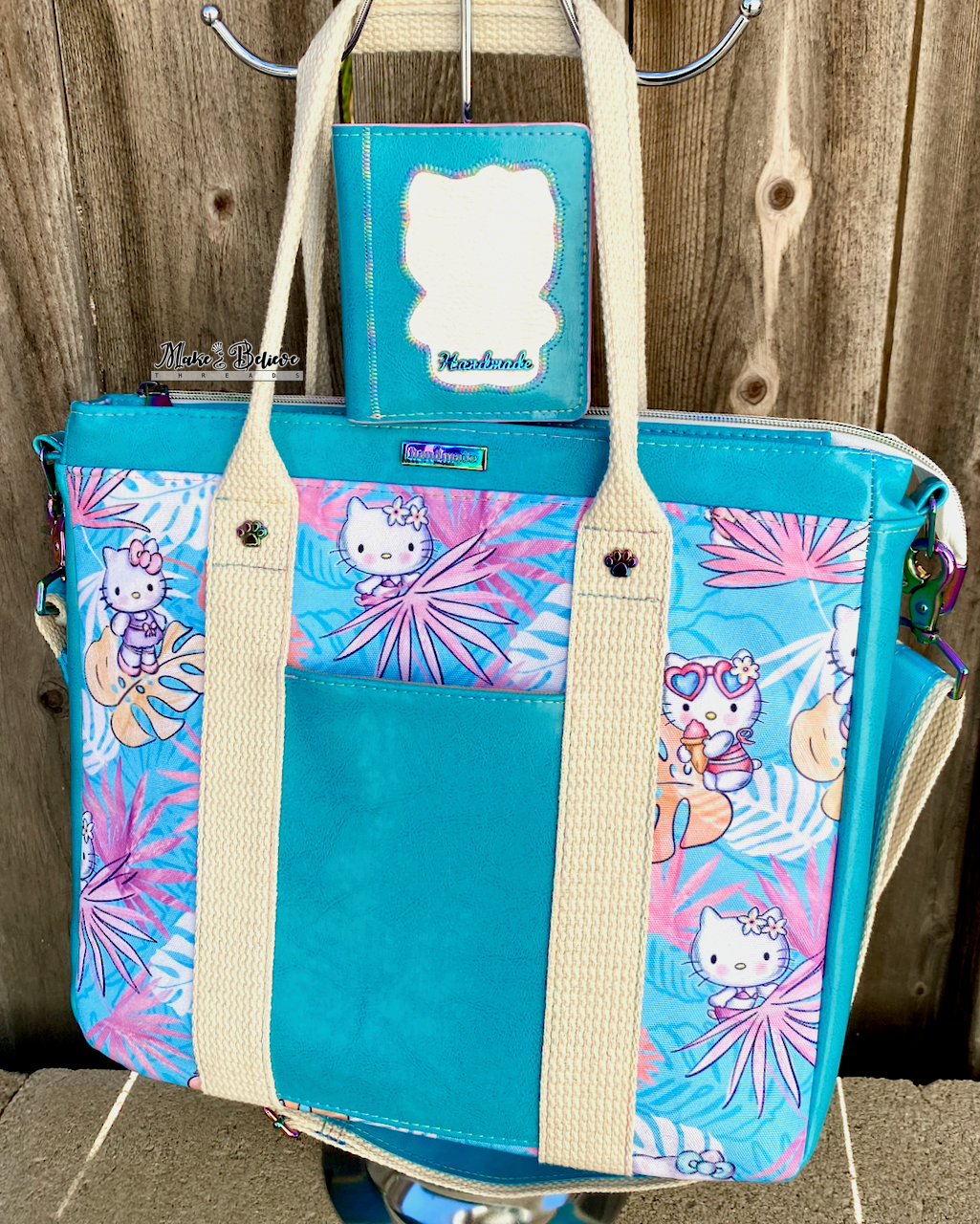 An aqua blue tote bag with pink and blue tropical print featuring "Bow Kitty" with a matching wallet sitting on top, photographed in front of a wooden fence.