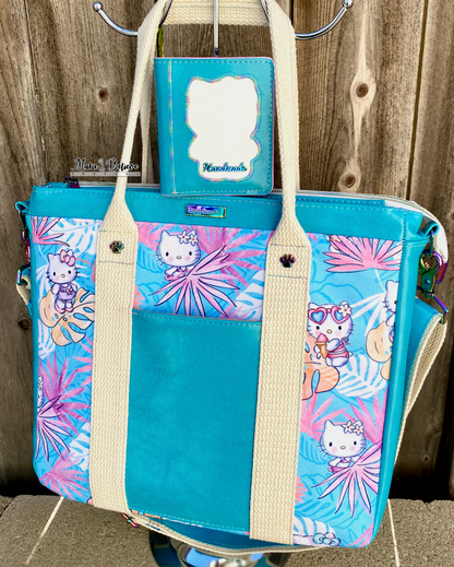 An aqua blue tote bag with pink and blue tropical print featuring "Bow Kitty" with a matching wallet sitting on top, photographed in front of a wooden fence.