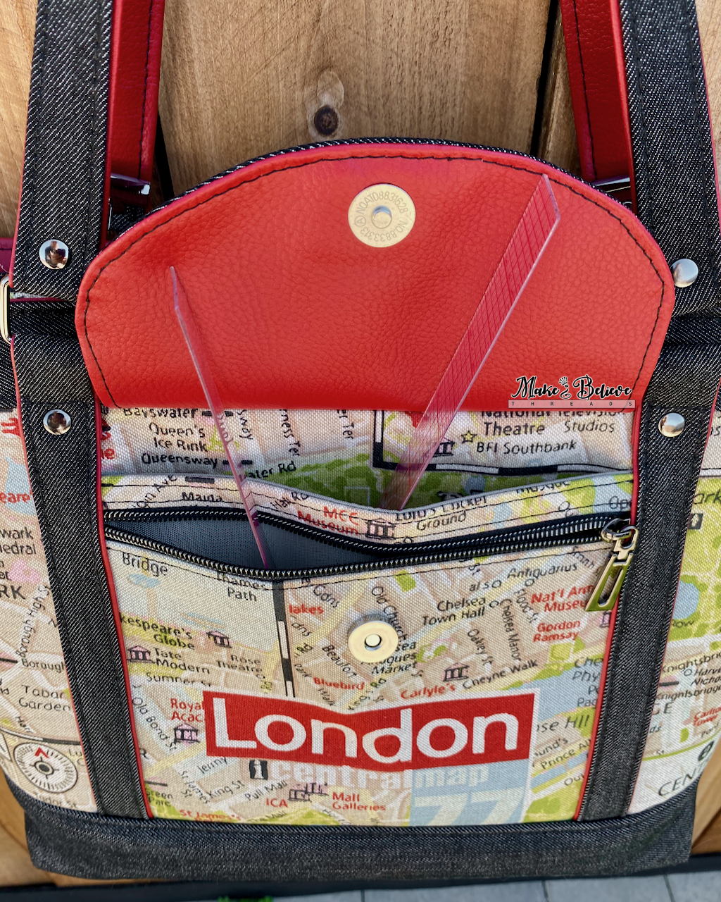 Black denim tote with red accents and map-pattern with 'London' text on a wooden surface. Photo illustrates an open front flap pocket revealing a slip pocket and zipper pocket.