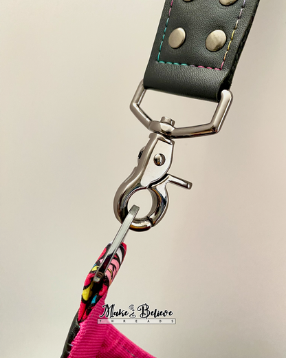 Close-up of a faux leather strap with gunmetal clasp, rainbow stitching, and rivets on a neutral background.