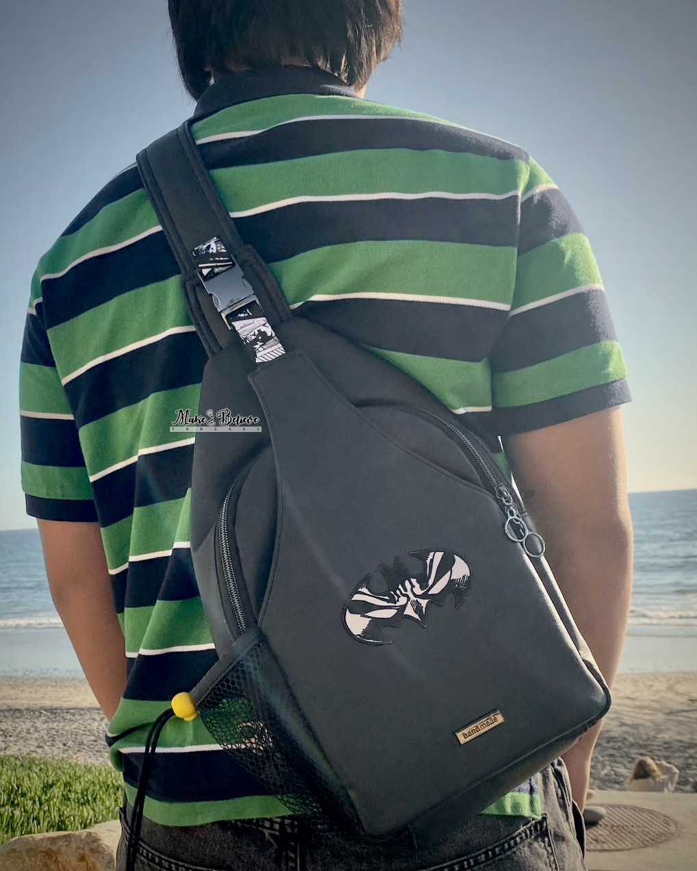 Person wearing a green and black striped shirt with a black faux leather sling bag with Batman patch and webbing on a beach.
