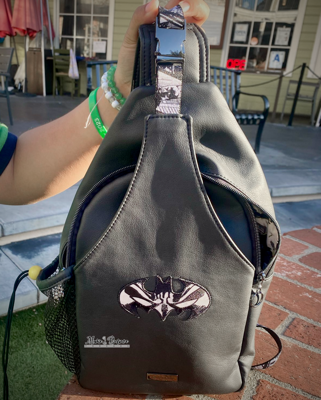 Black faux leather sling bag with a Batman patch and webbing held by a person outdoors.