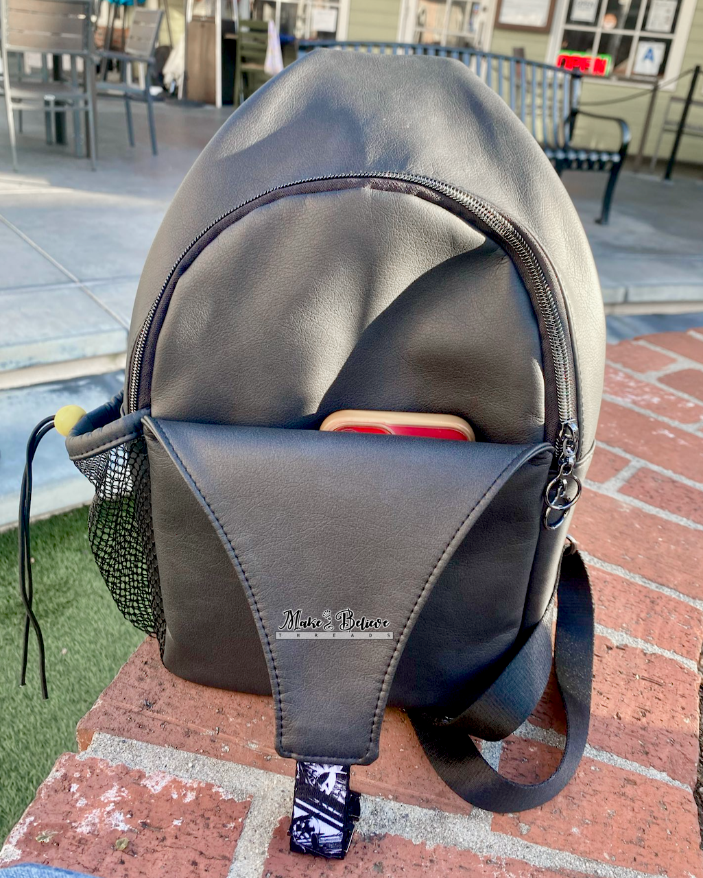 Black faux leather sling bag with a small mesh side pocket on a red brick surface.