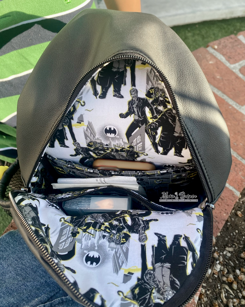 Black faux leather sling bag interior with Gotham villains design, featuring a padded slip pocket and zipper pocket, photographed on a person's lap outdoors.