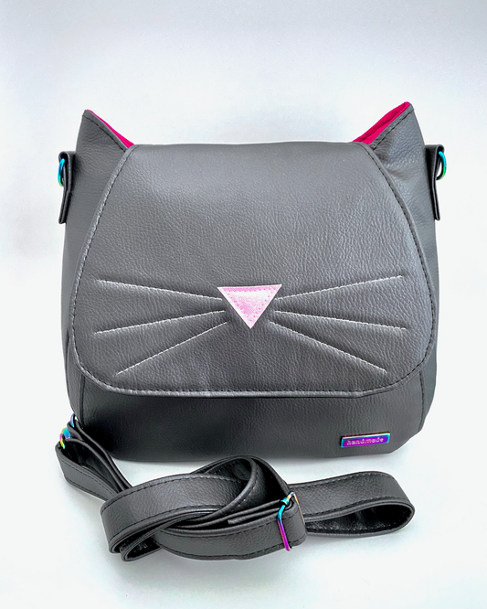 Gray crossbody bag shaped like a cat face with a pink nose, and wiskers on a white background