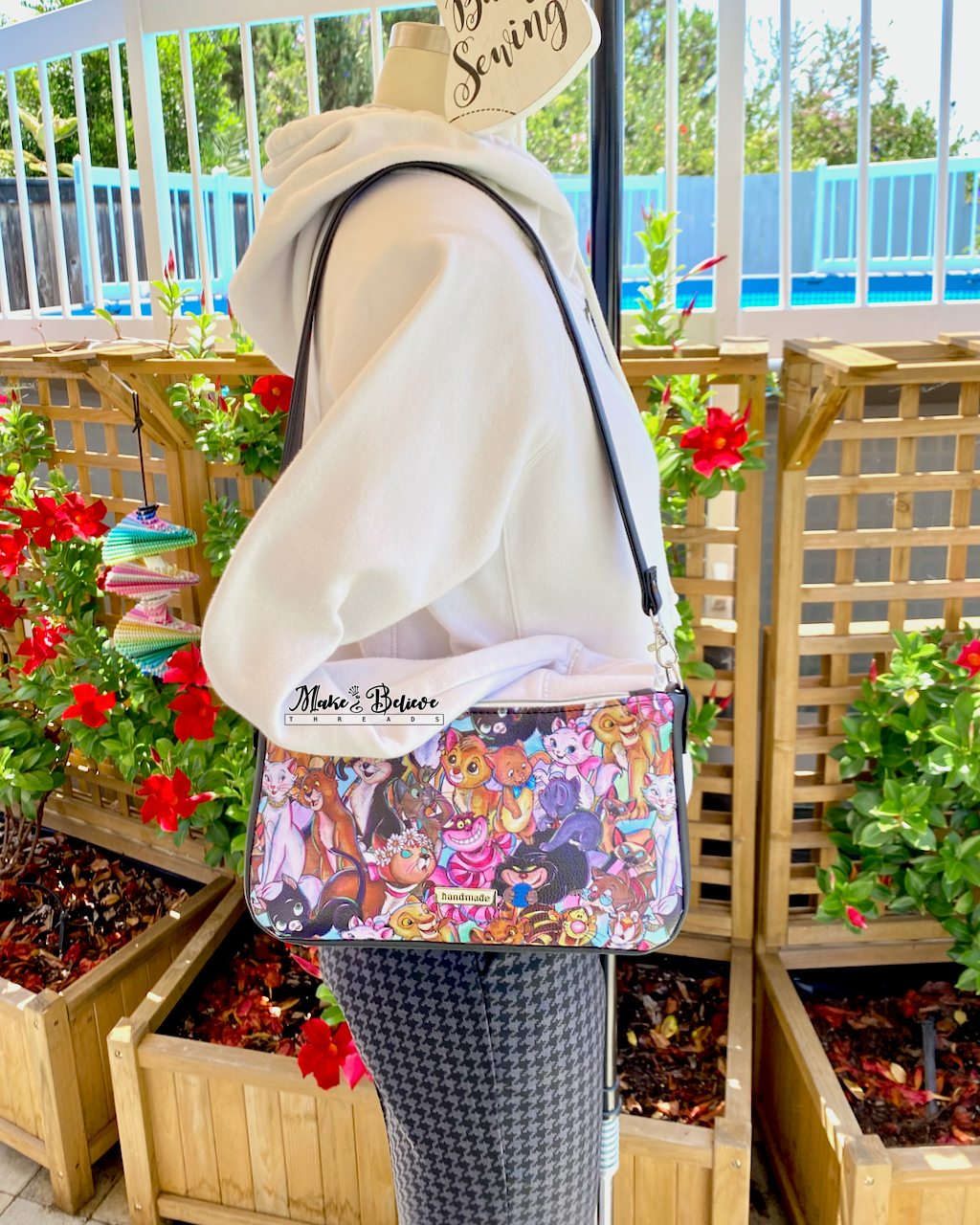 Mannequin wearing a white hoodie wearing a colorful cartoon cats shoulder bag in an outdoor setting with plants and flowers.