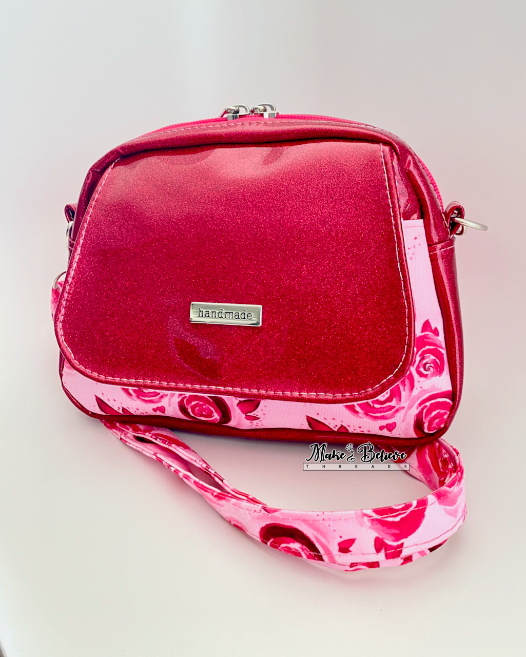 Pinkish-red glitter crossbody bag with floral  pockets and strap on a light gray background