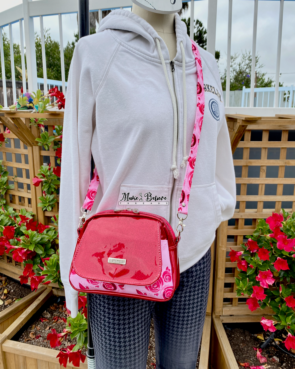 Mannequin wearing a white hoodie and a Pinkish-red glitter crossbody bag with floral  pockets and strap in an outdoor setting with flowers and wooden lattice.