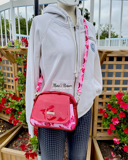 Mannequin wearing a white hoodie and a Pinkish-red glitter crossbody bag with floral  pockets and strap in an outdoor setting with flowers and wooden lattice.