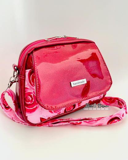 Front flap over slip pocket pocket on pinkish-red glitter crossbody bag with rose pattern on a white background