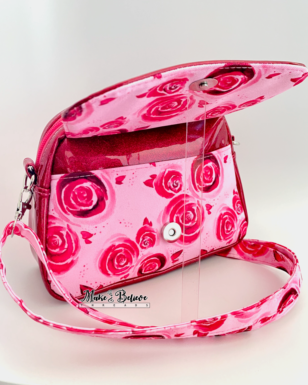 Open front flap revealing slip pocket pocket on pinkish-red glitter crossbody bag with rose pattern on a white background