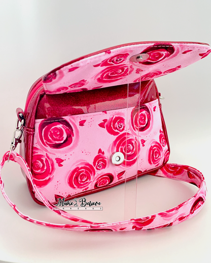 Open front flap revealing slip pocket pocket on pinkish-red glitter crossbody bag with rose pattern on a white background