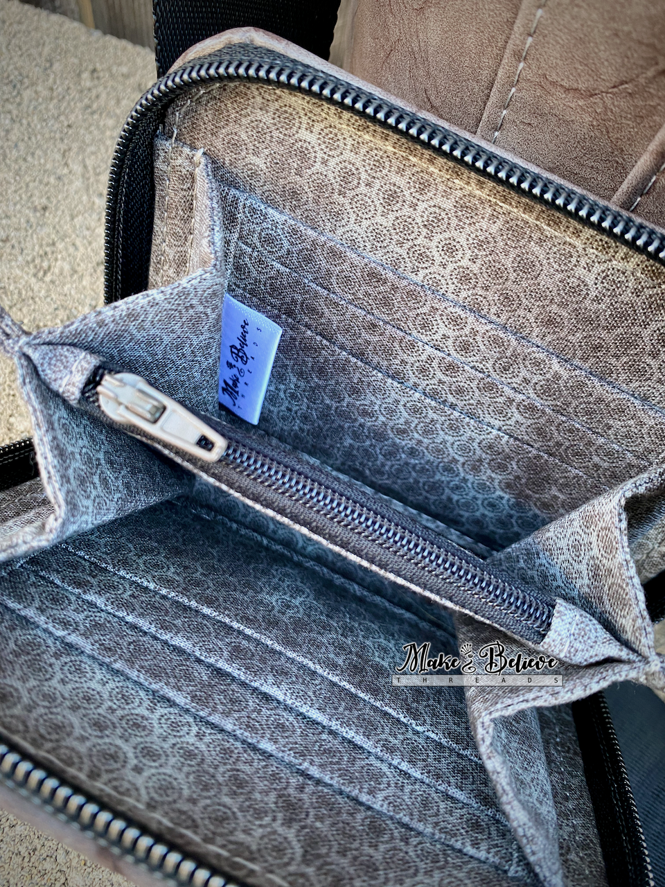Brownish-gray zip-around wallet with zipper and patterned interior, featuring a Make Believe Threads woven label.