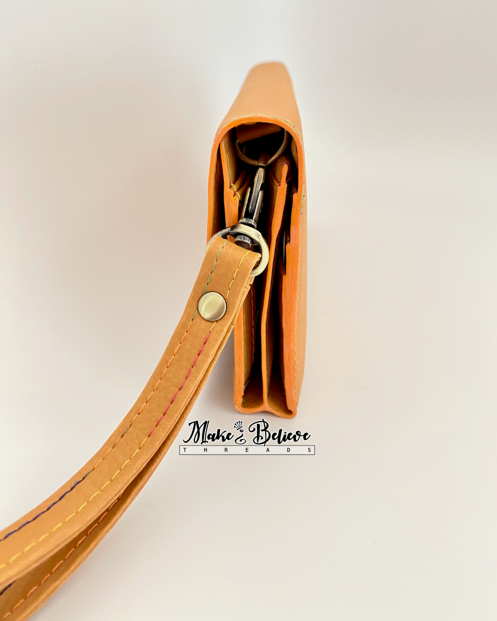 Side view of ochre tri-fold wallet with strap on a neutral background, featuring coordinating pumpkin-colored edge painting and antique brass hardware