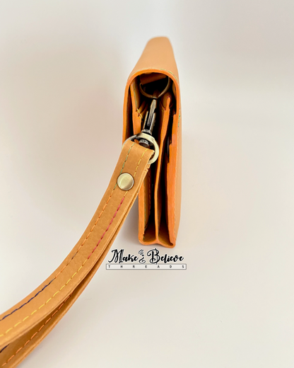 Side view of ochre tri-fold wallet with strap on a neutral background, featuring coordinating pumpkin-colored edge painting and antique brass hardware