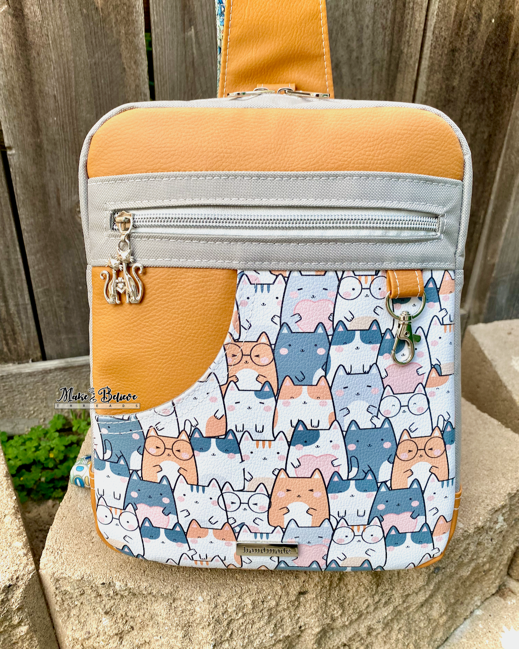 Medium-sized rectangular sling bag with cat pattern and orange top section on a stone surface.
