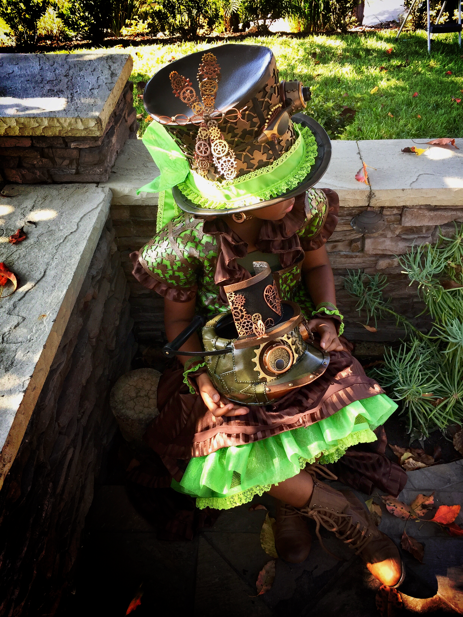 A young Black female in elaborately handcrafted steampunk Princess Tiana costume with large hat and brown dress with green accents outdoors. She is holding a trick-or-treat bag shaped as a steampunk frog head.