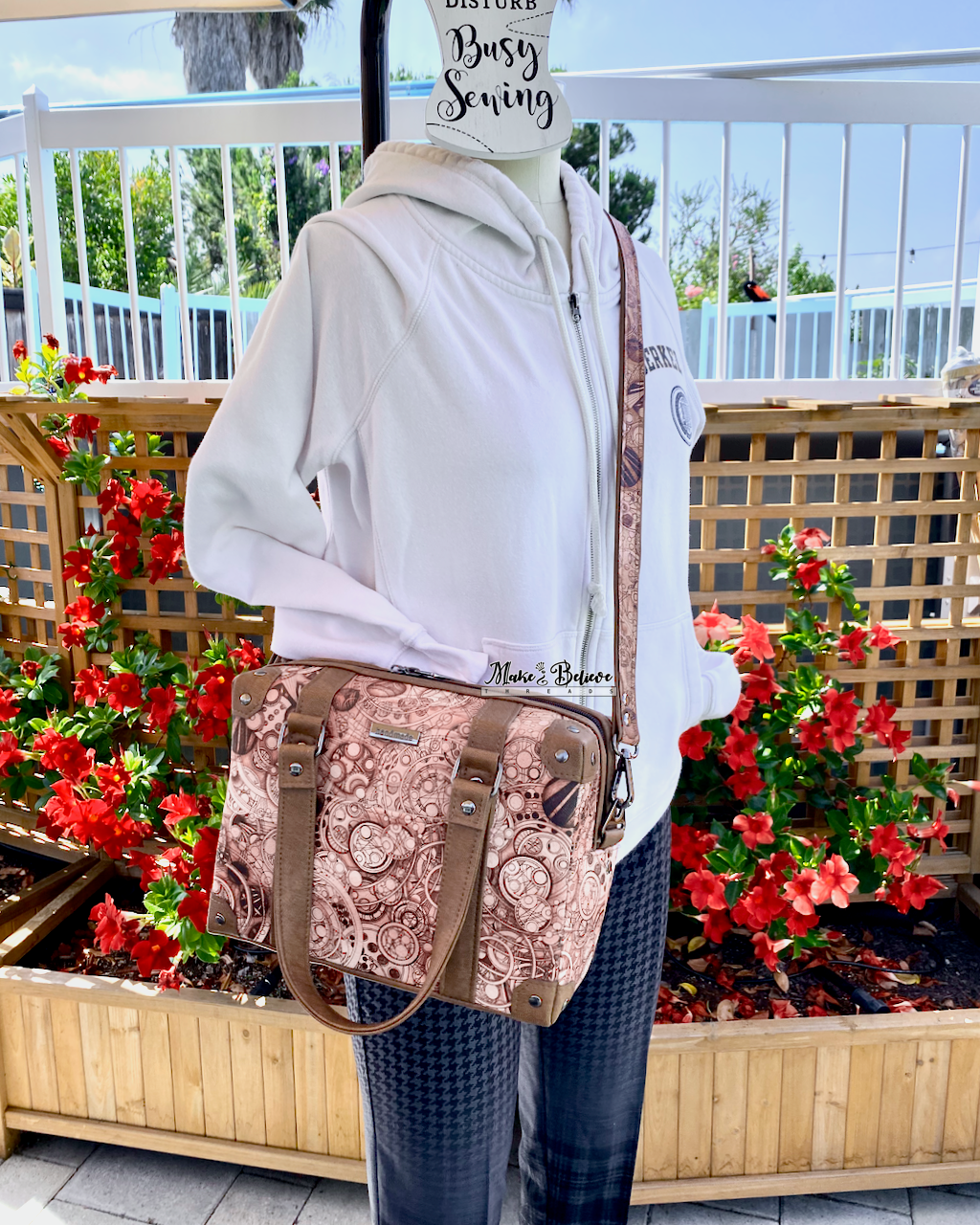 Mannequin with a white hoodie, wearing a steam-trunk shaped handbag with brown accents as a crossbody bag, in front of red flowers.